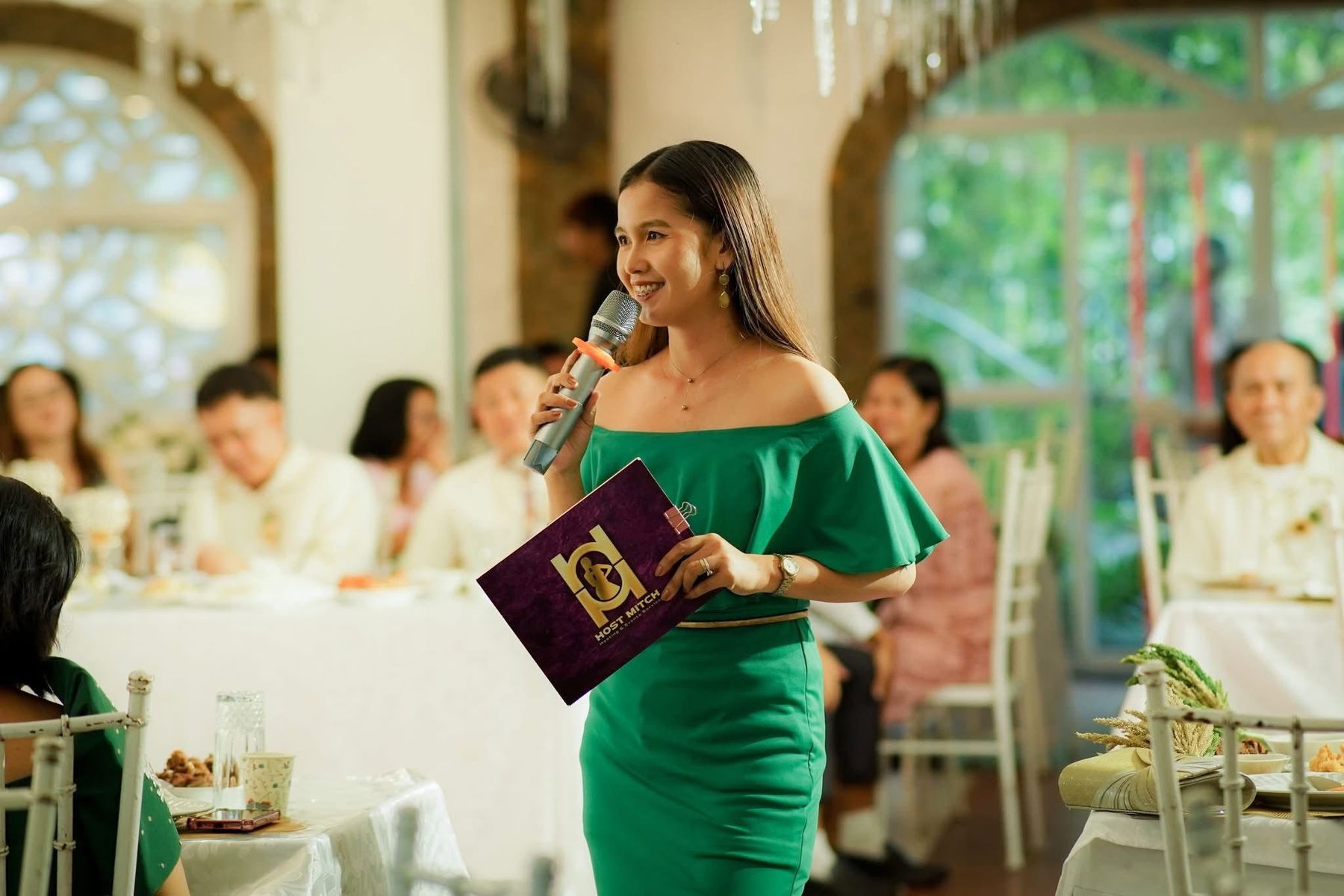 Host Mitch holding a microphone and her program folder, speaking to guests at a reception
