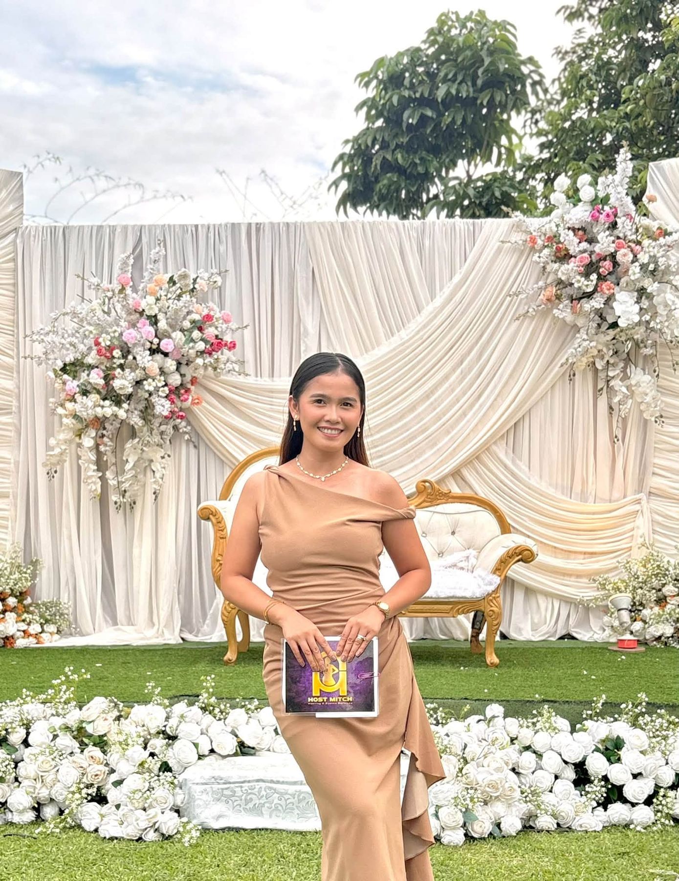 Host Mitch smiling in a garden venue, holding her program folder